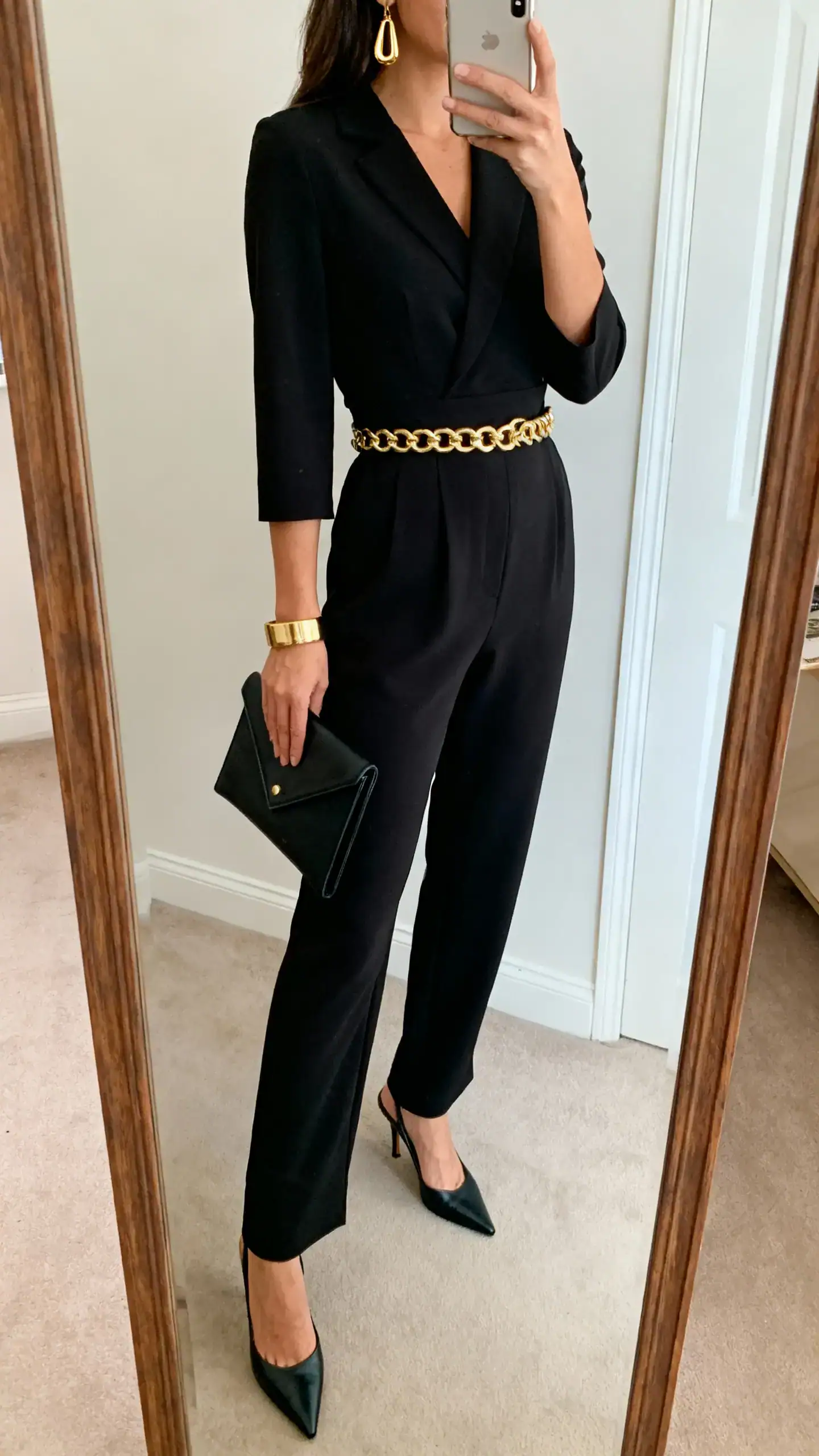 iPhone side-angle mirror selfie of a woman wearing a tailored black jumpsuit with a defined waist, gold chain belt, pointed-toe heels, gold drop earrings, cuff bracelet, and a mini envelope clutch; face not visible, full-length mirror, soft indoor lighting, iPhone photo quality.